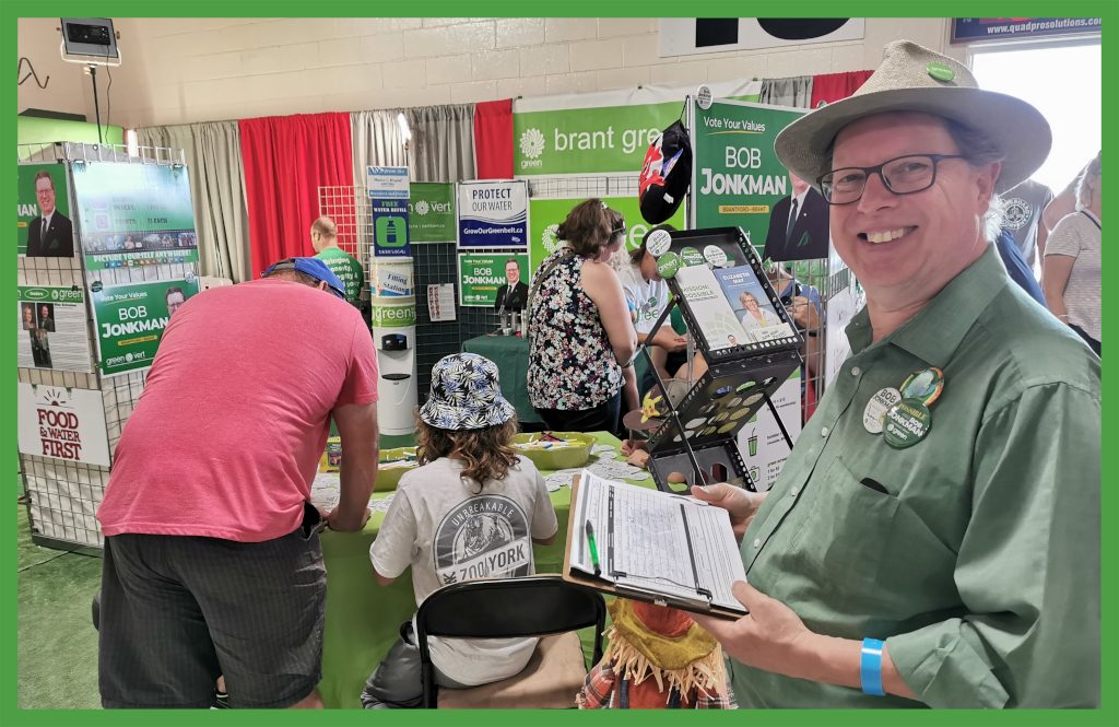 Bob stands at the entrance to the Brant Greens Paris Fair booth. Behind him you can see people making buttons and part of the photo booth. 